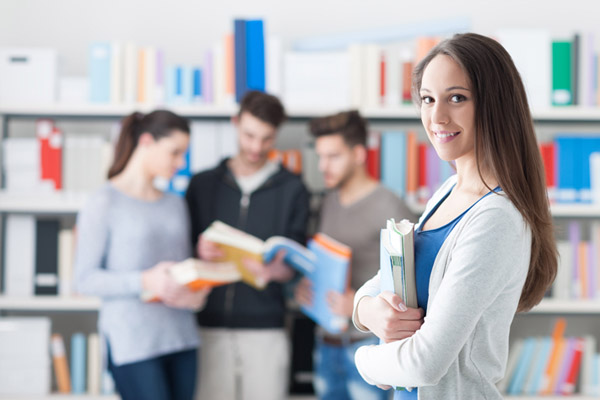 Mujer con sueter blanco y blusa azul abrazando un libro y al fondo tres personas con libros