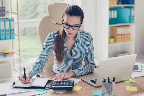 Mujer con blusa negra rayada sentada frente a un escritorio con una laptop, calculadora y libreta