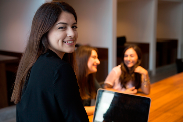 Mujer con saco negro sentada frente una laptop y al fondo otras dos mujeres sentadas