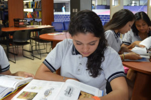 Mujer con playera gris sentada leyendo un libro dentro de una biblioteca de la preparatoria Acapulco