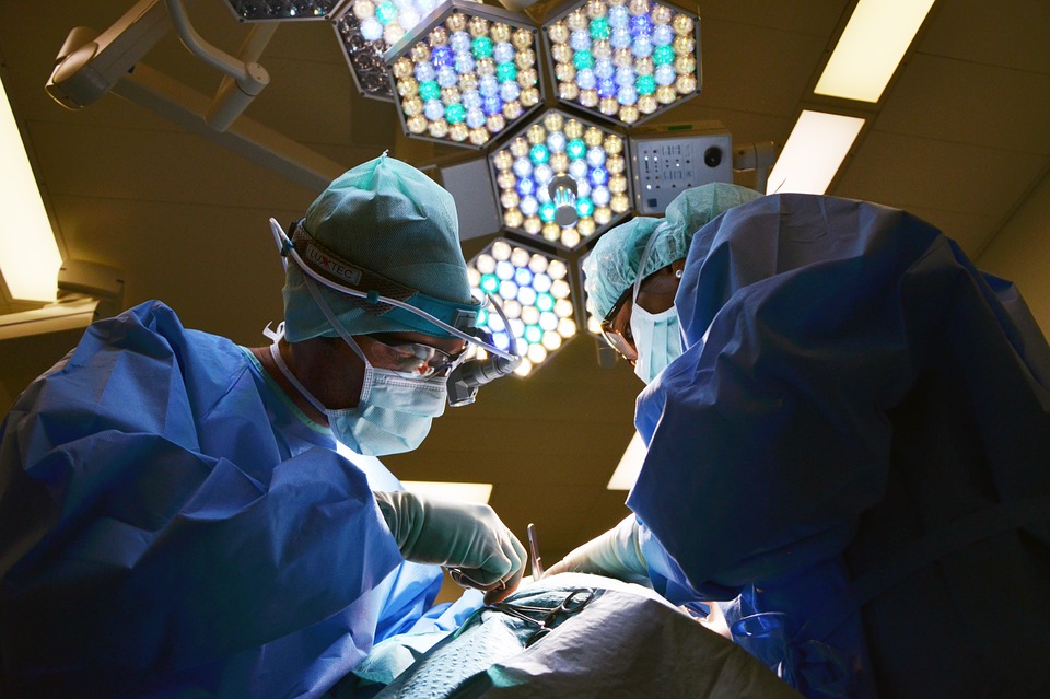 Hombre y mujer con bata azul, cubrebocas , lentes y gorro bajo una lámpara realizando una operación