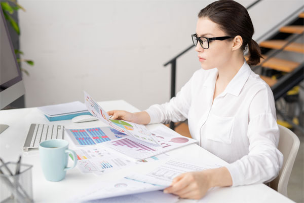Mujer con blusa blanca sentada frente a un escritorio revisando papeles con gráficas